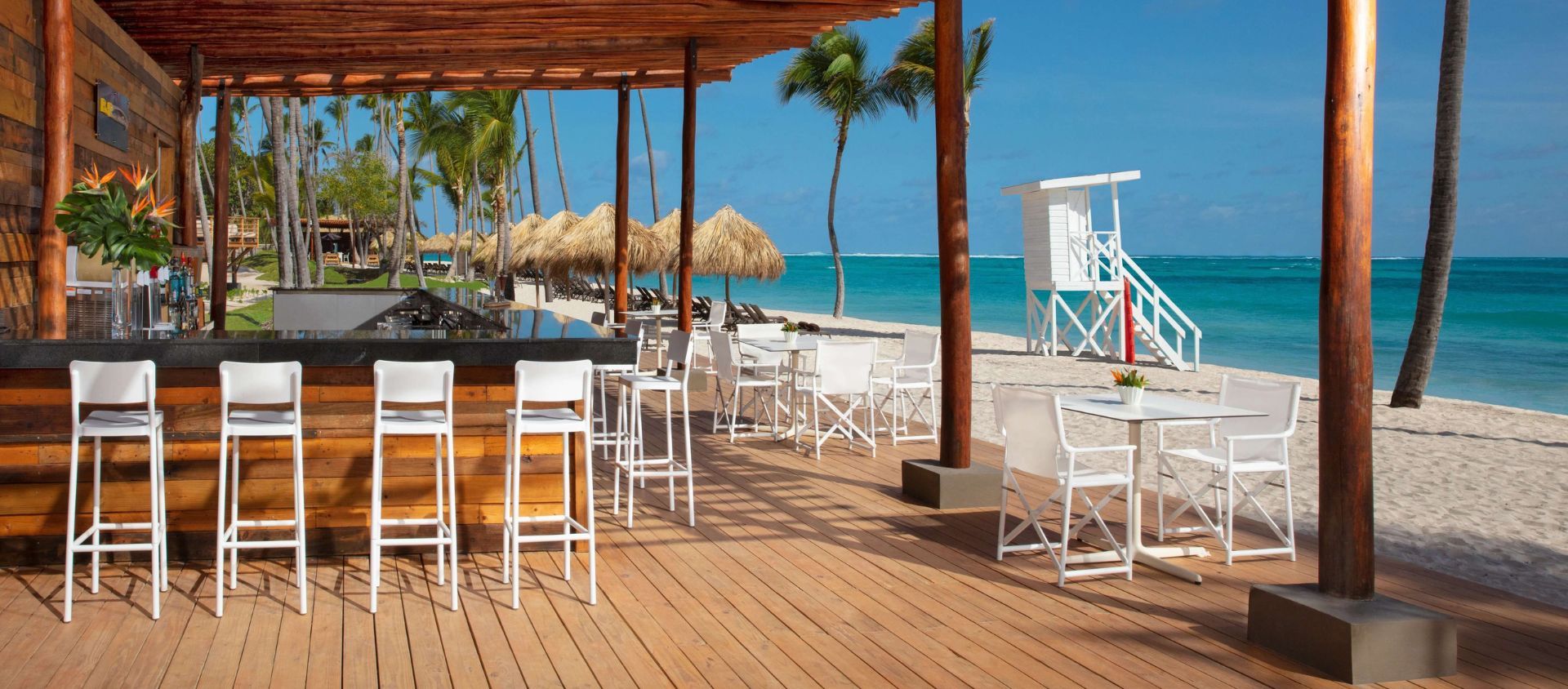 Beachfront bar with white stools and oceanfront seating under a wooden pavilion.