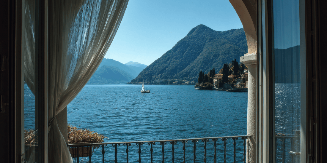 View through an open window of a blue lake with mountains and a sailboat.