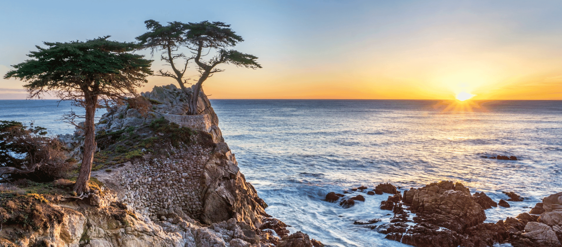 Rocky coastal cliff at Carmel-by-the-sea with windswept trees overlooking the ocean at sunset.