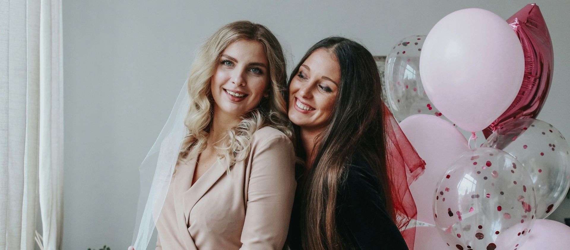 A bride in a veil poses with a friend beside pink balloons.