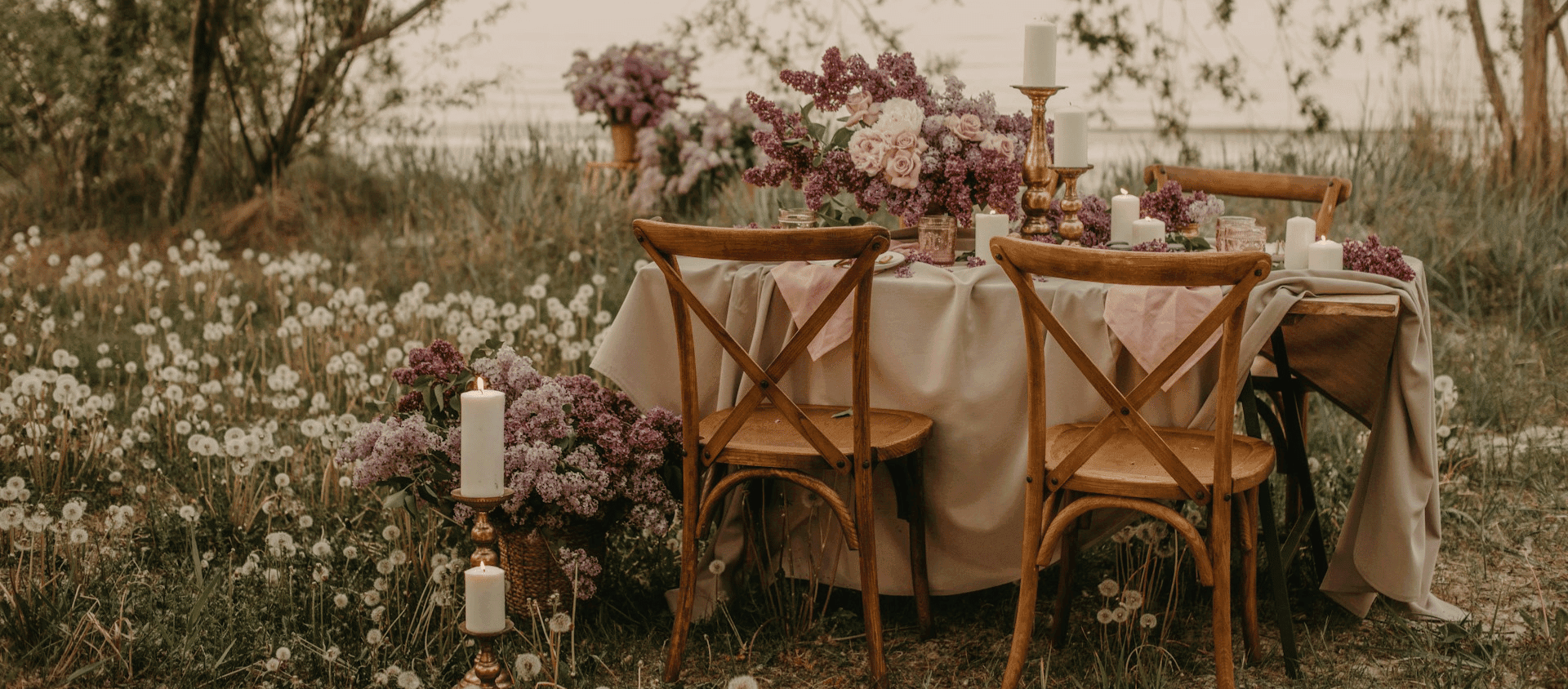 Rustic outdoor table with candles, lilac flowers, and wooden chairs in a field setting.