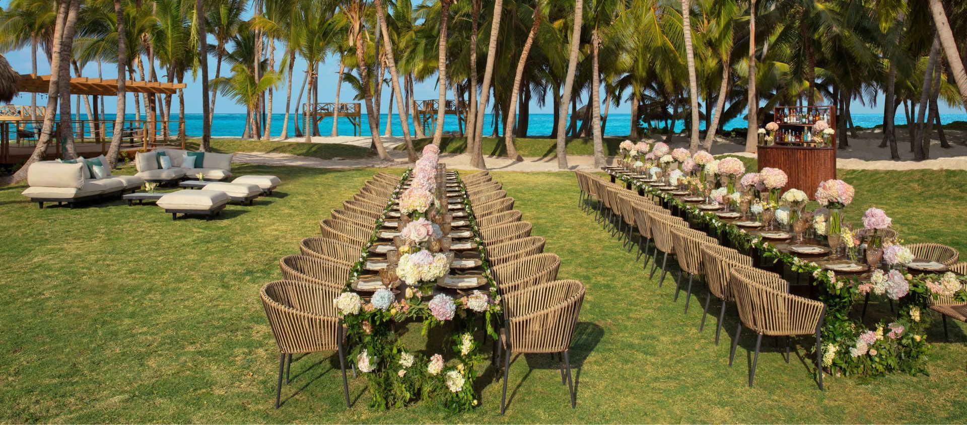 Long banquet tables with floral runners set on a beachfront lawn lined with palm trees.