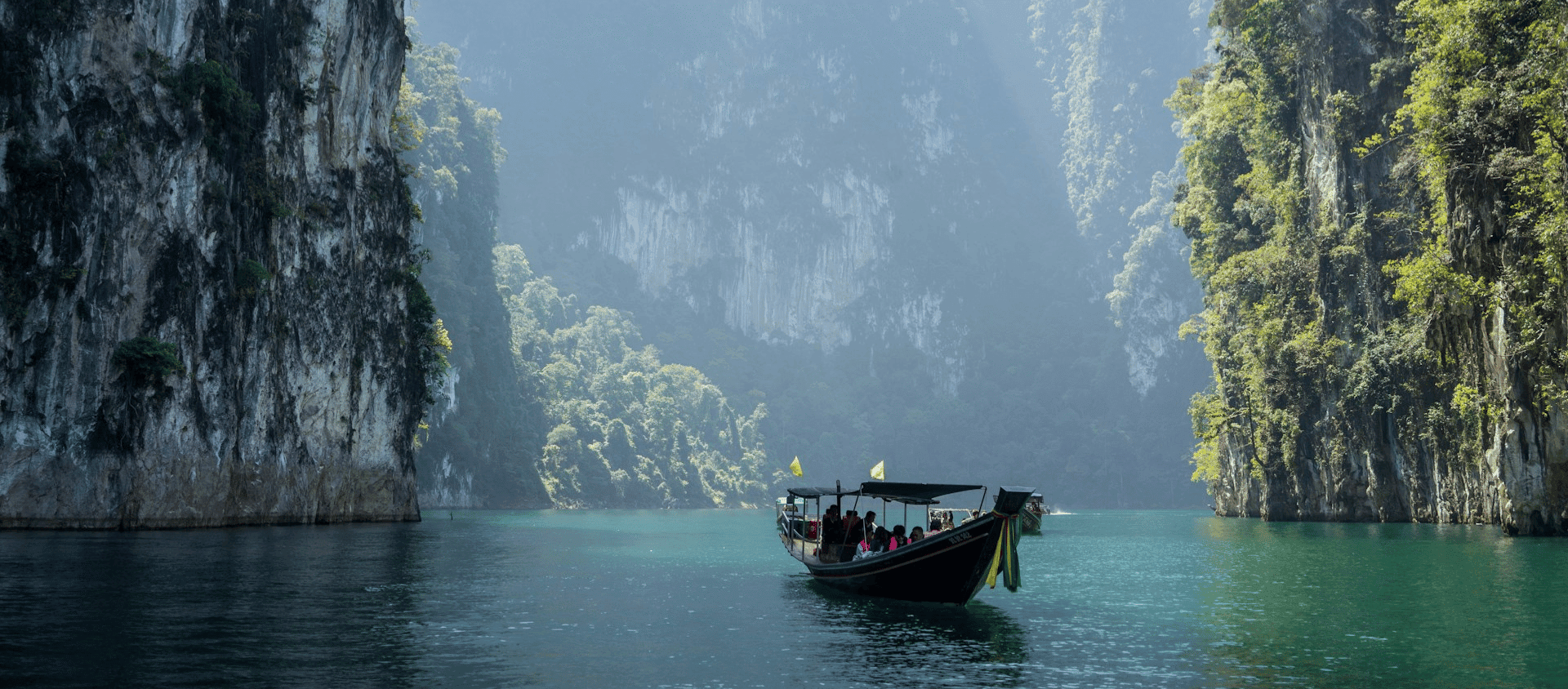 A boat carrying passengers on turquoise water between tall, forested cliff in Thailand