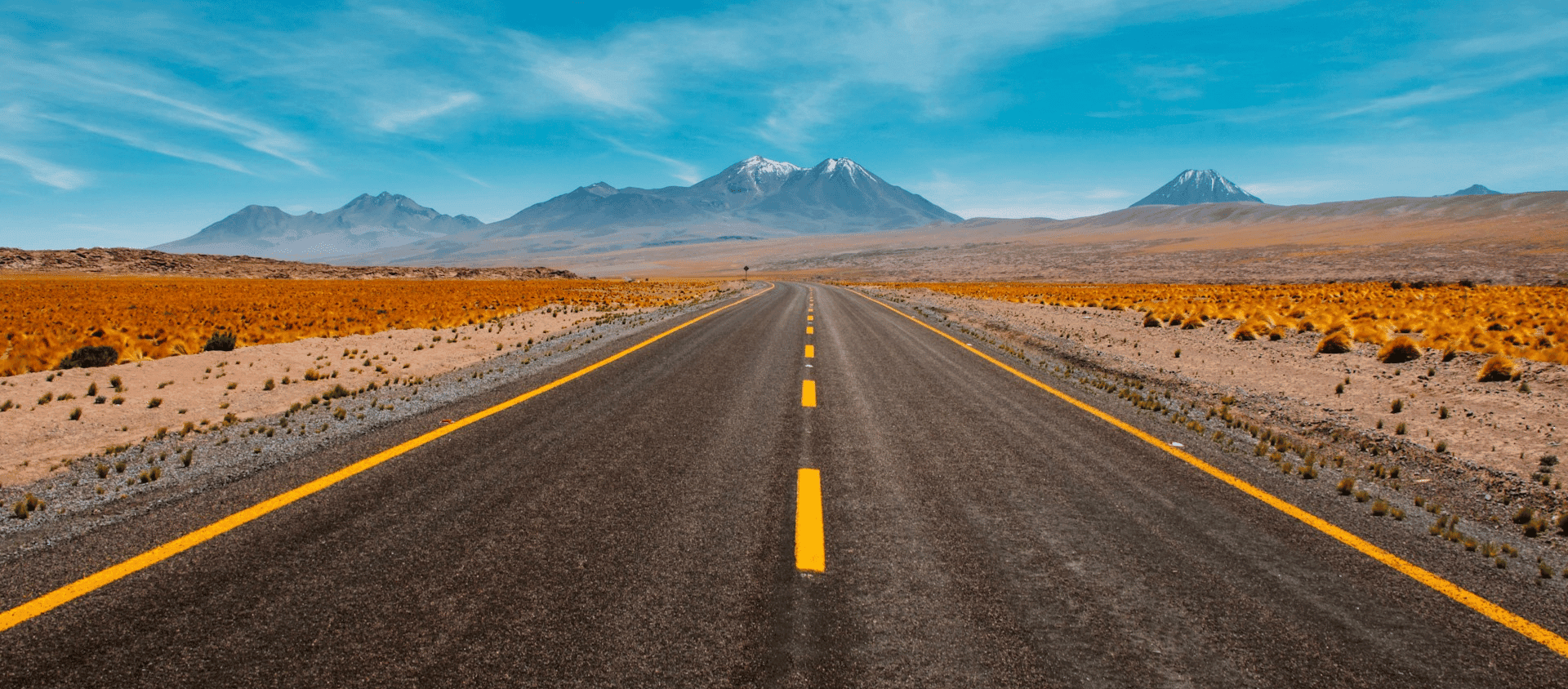 Long straight highway through a desert landscape leading toward distant snow-capped mountains under a blue sky.