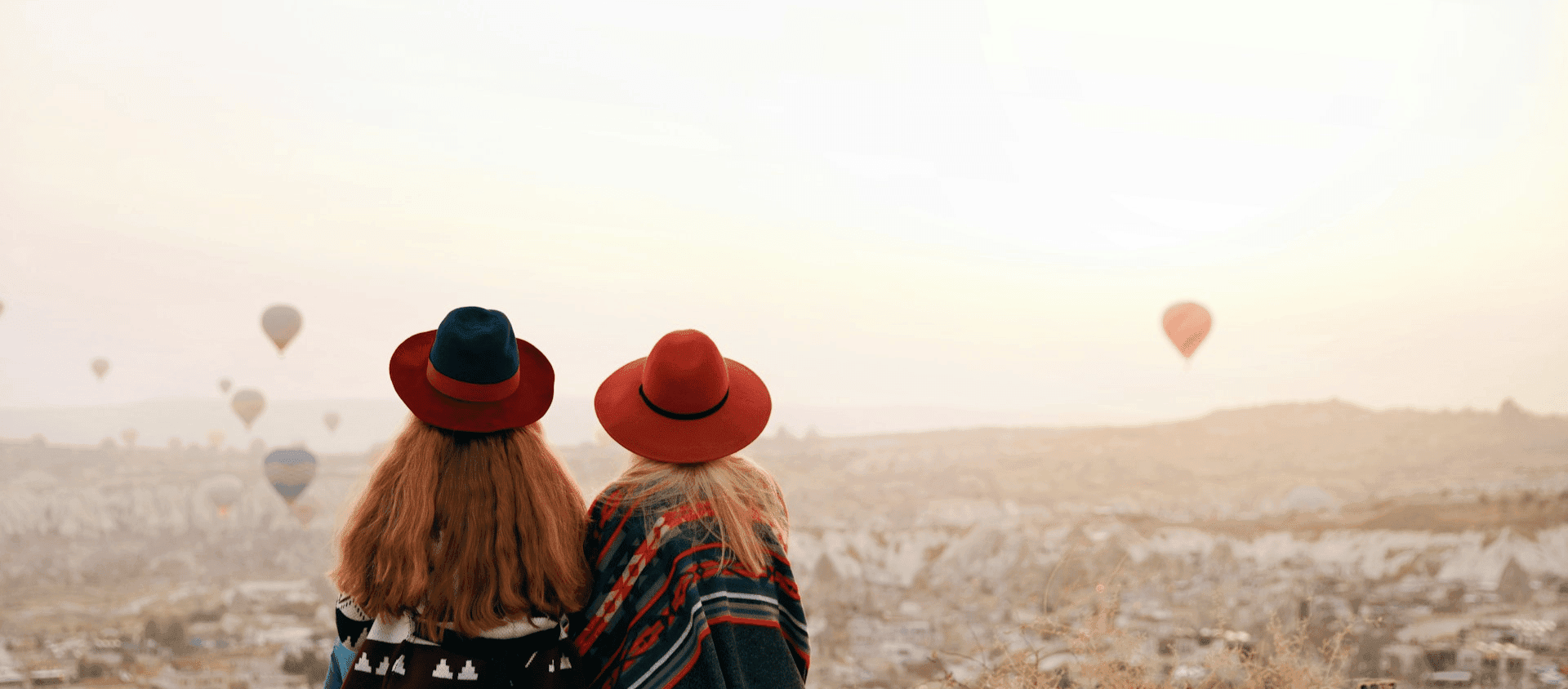 Two women sitting together watching colorful hot air balloons float over a desert landscape.