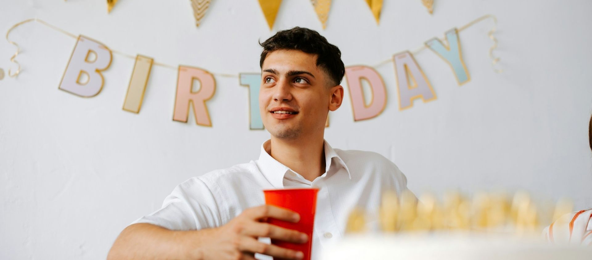 Young man holding a red cup at a birthday party with a colorful banner behind him.