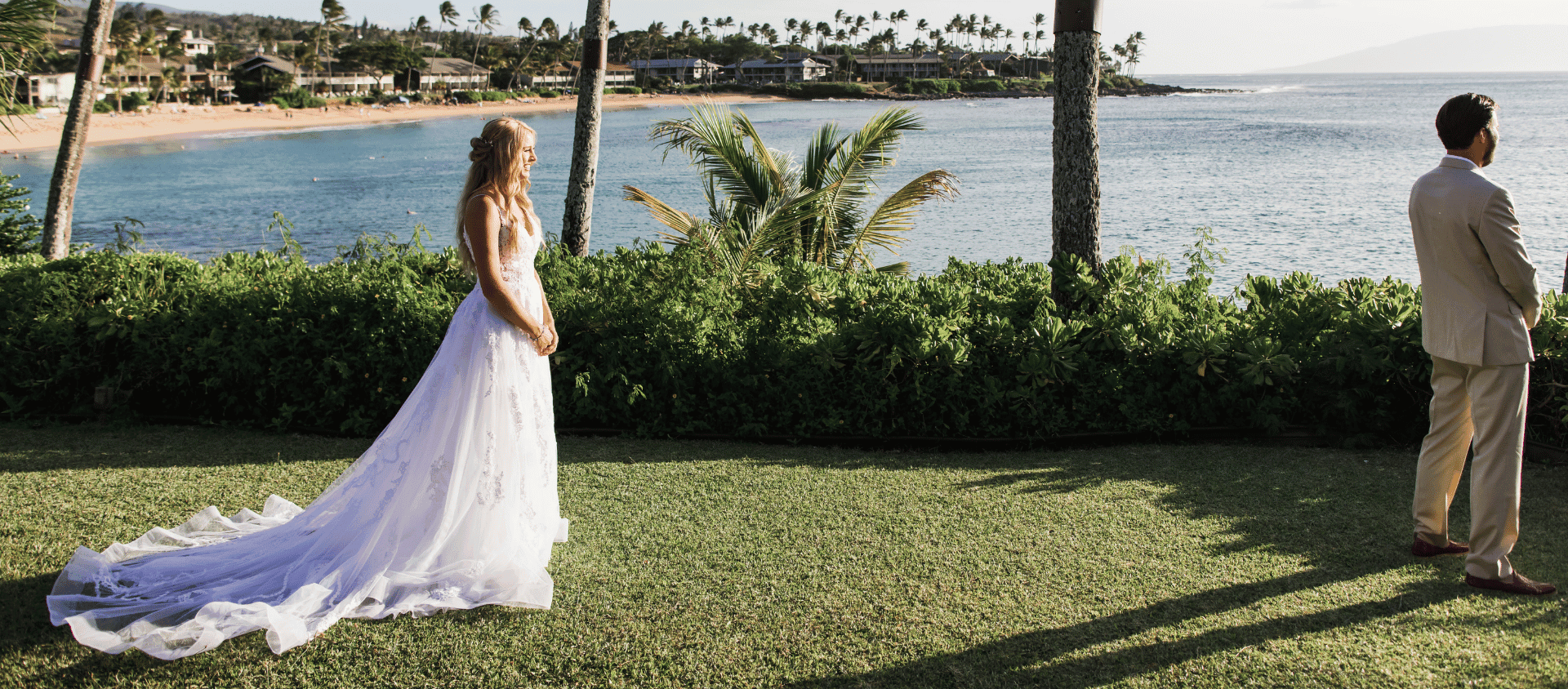 Bride and groom standing on lawn at Napili Kai Beach Resort overlooking ocean at sunset.
