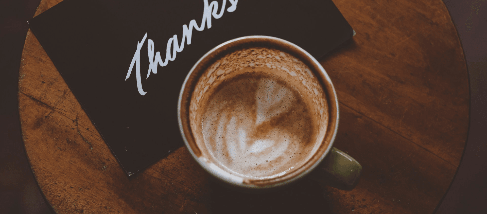 Cup of coffee on a wooden table beside a black card that reads “Thanks!”