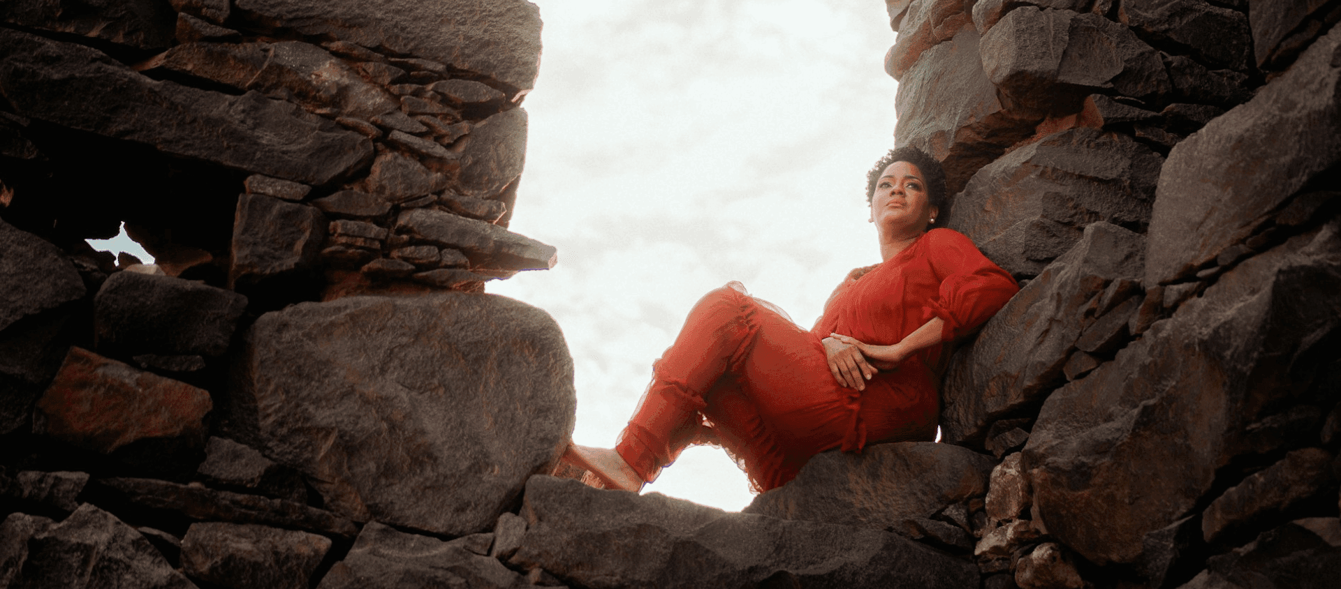 Woman in a red outfit sitting in a dramatic rocky coastal landscape.