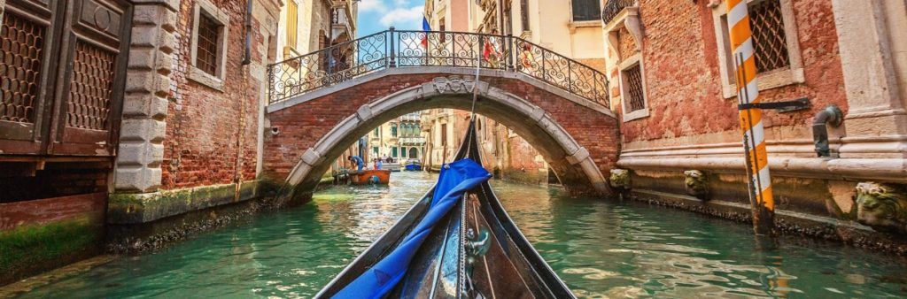 View from a gondola moving through a Venice canal toward a small arched bridge between historic buildings.