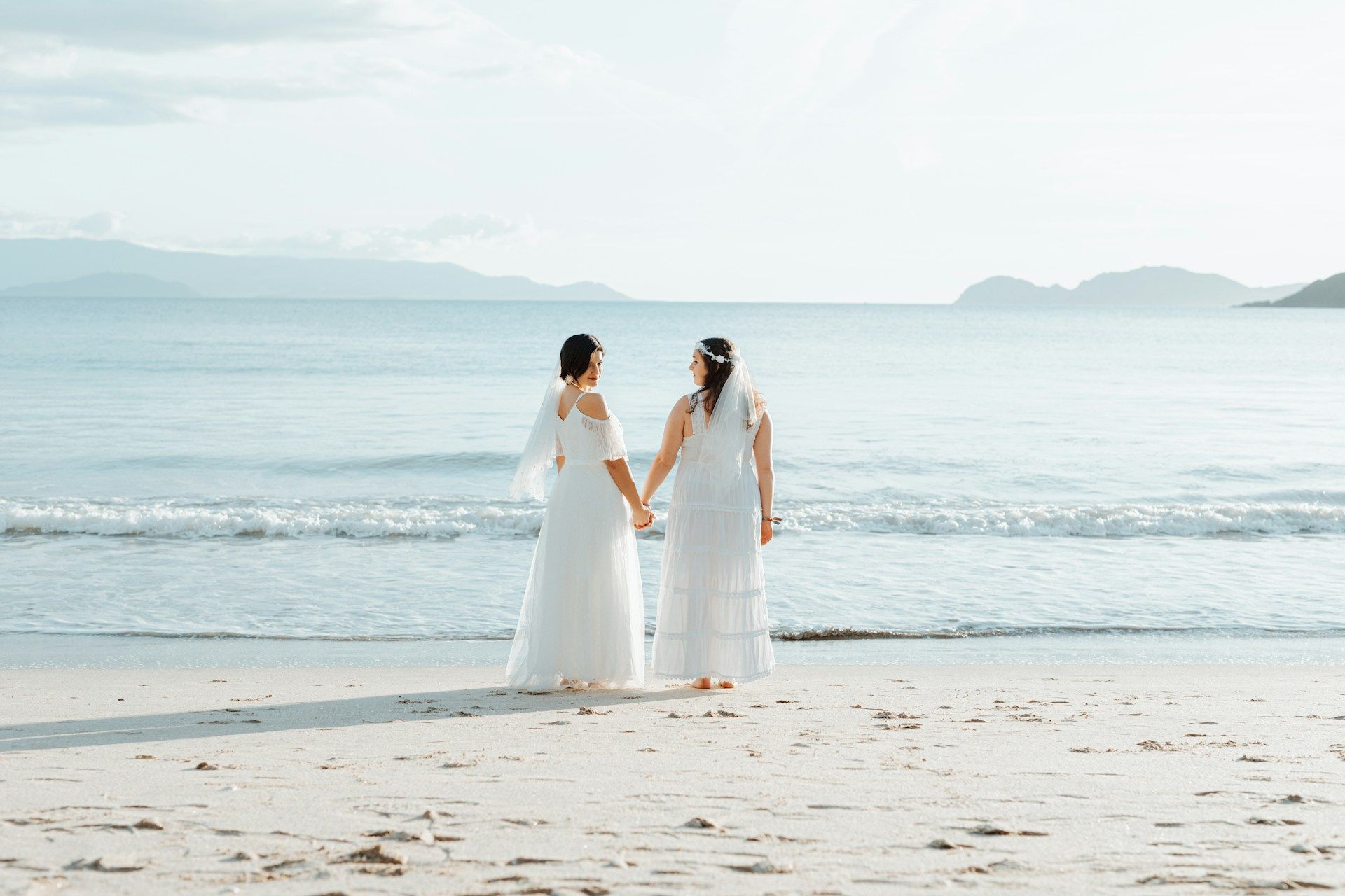 two brides holding hands on shoreline with ocean in background