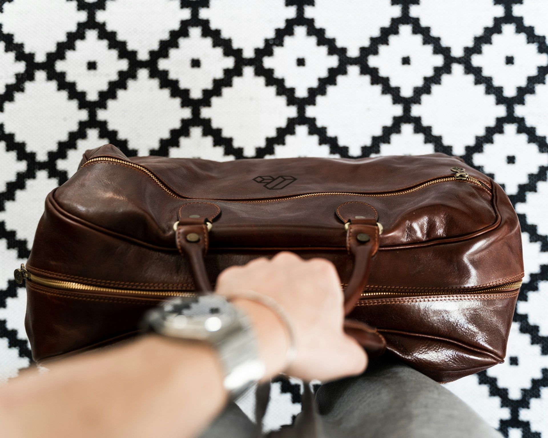 Hand holding a brown leather duffel bag against a black and white patterned background.