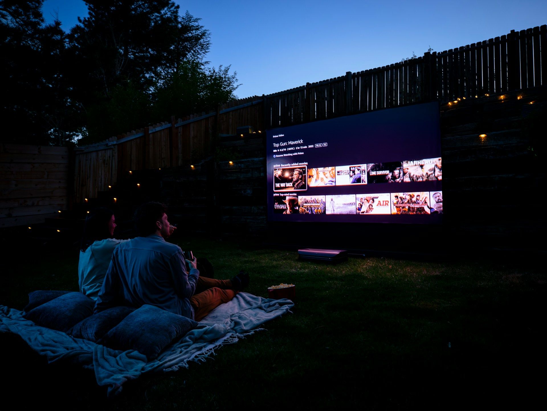 Couple watching a movie on an outdoor projector screen in a backyard at night.