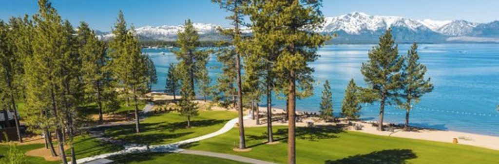 Scenic view of Lake Tahoe with pine trees, green lawns, and snow-capped mountains in the distance.