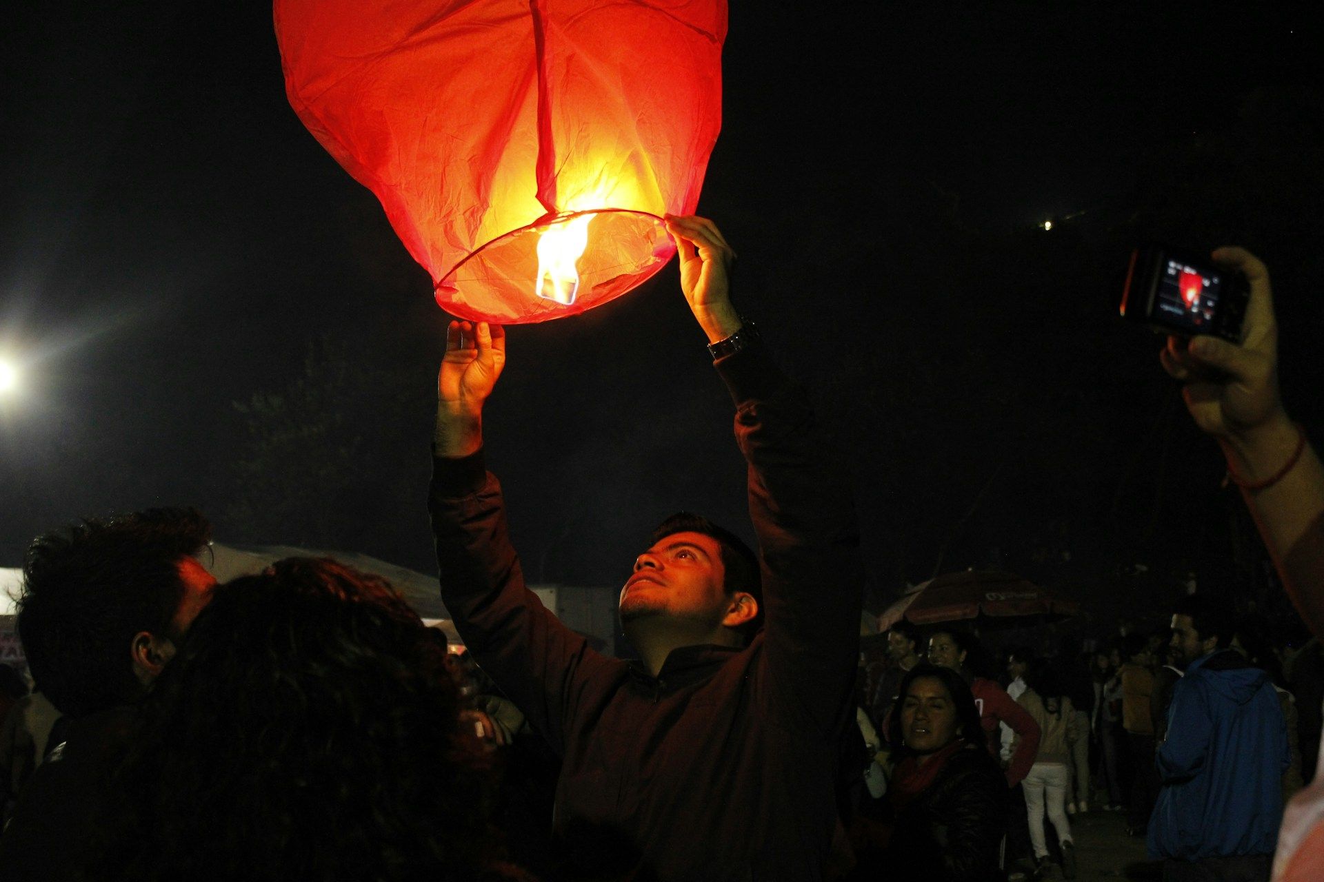 Person releasing a glowing red paper lantern into the night sky at an outdoor gathering