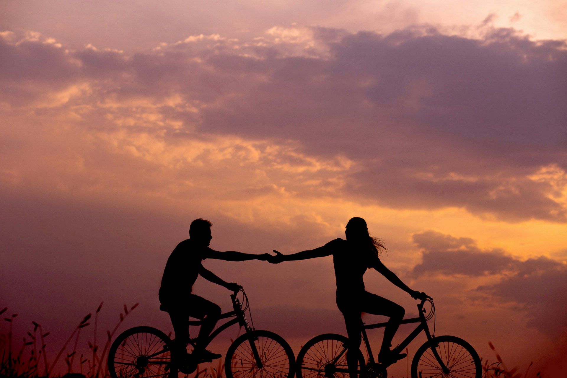 Silhouette of two people riding bicycles at sunset, reaching toward each other.