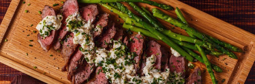 Sliced steak topped with creamy sauce and herbs, served with asparagus on a wooden board.