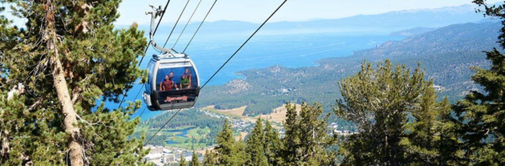 Gondola lift ascending over forested hills with a view of Lake Tahoe in the background.