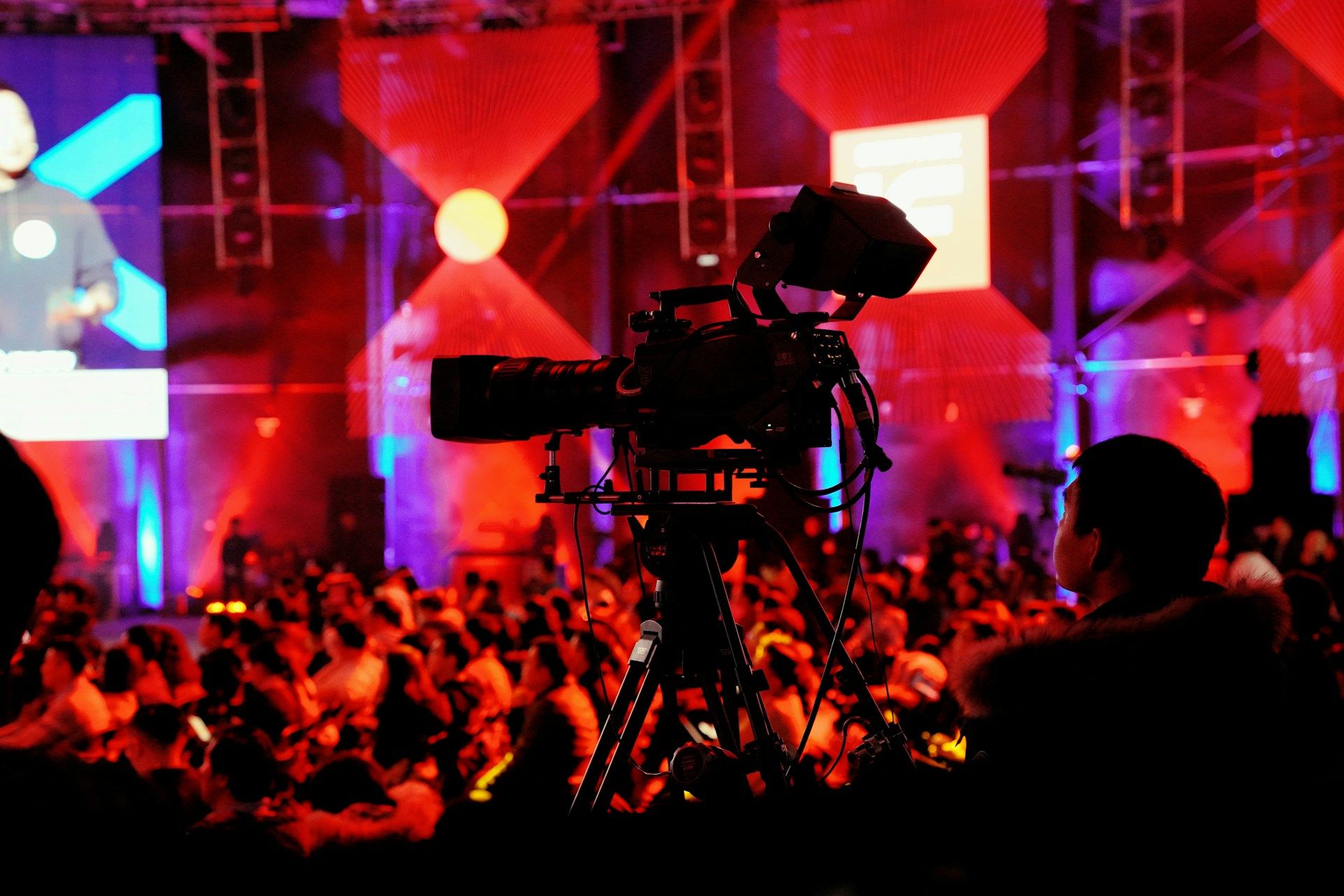 Video camera on a tripod filming a live event with a large audience and red stage lighting.