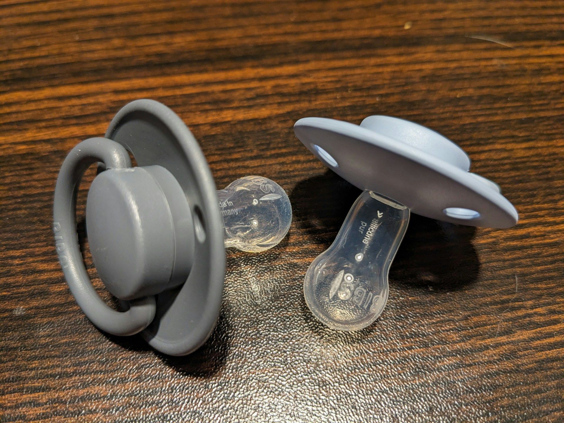 Two gray silicone baby pacifiers on a wooden surface.