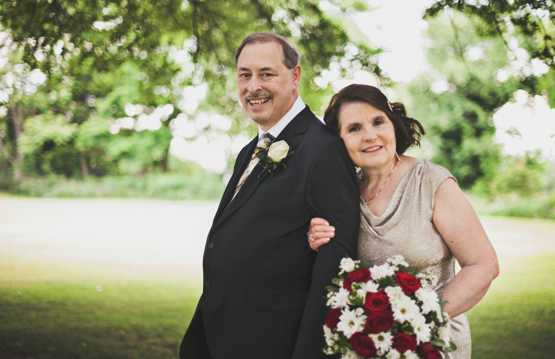 Formally dressed couple smiling outdoors, woman holding a red and white bouquet.