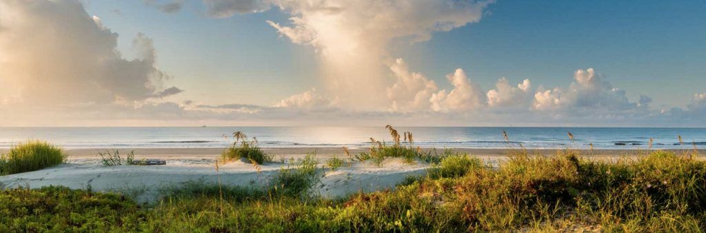 Peaceful beach at Kiawah Island, South Carolina with grassy dunes, soft sand, and calm ocean waves under a partly cloudy sky.