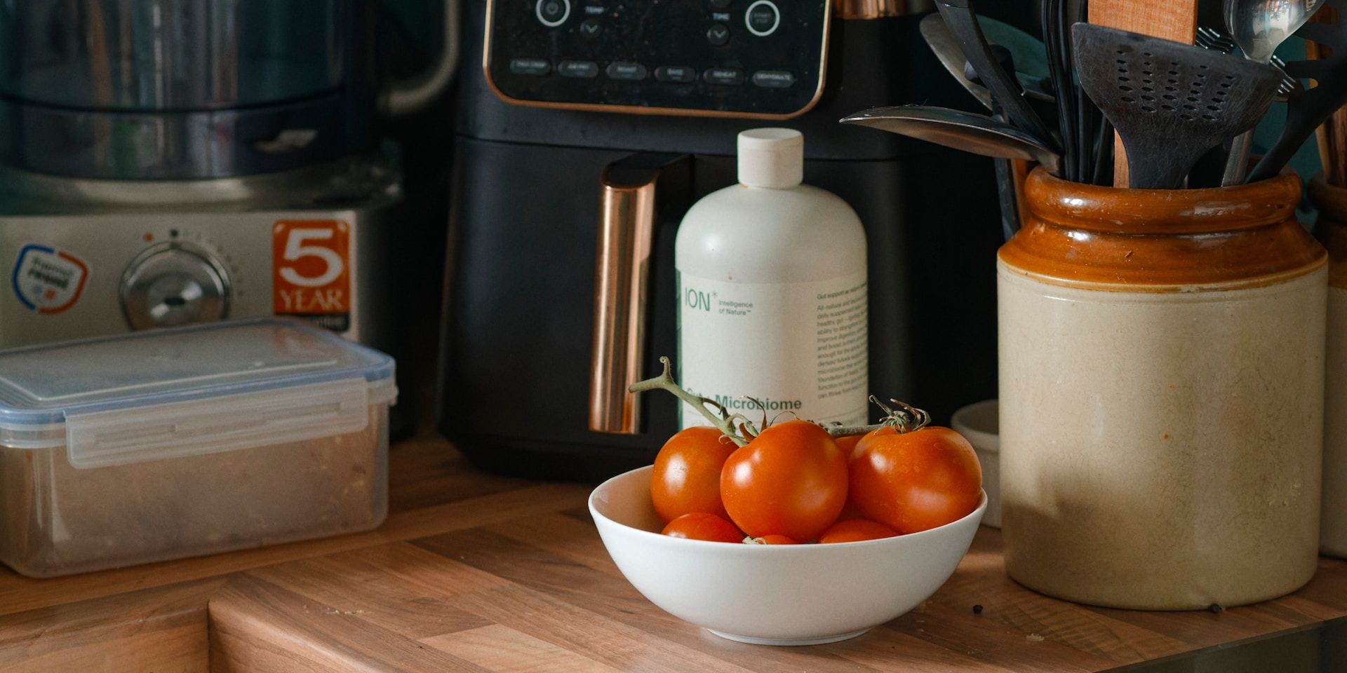 Air fryer on a wooden kitchen counter beside a bowl of fresh tomatoes and cooking utensils.
