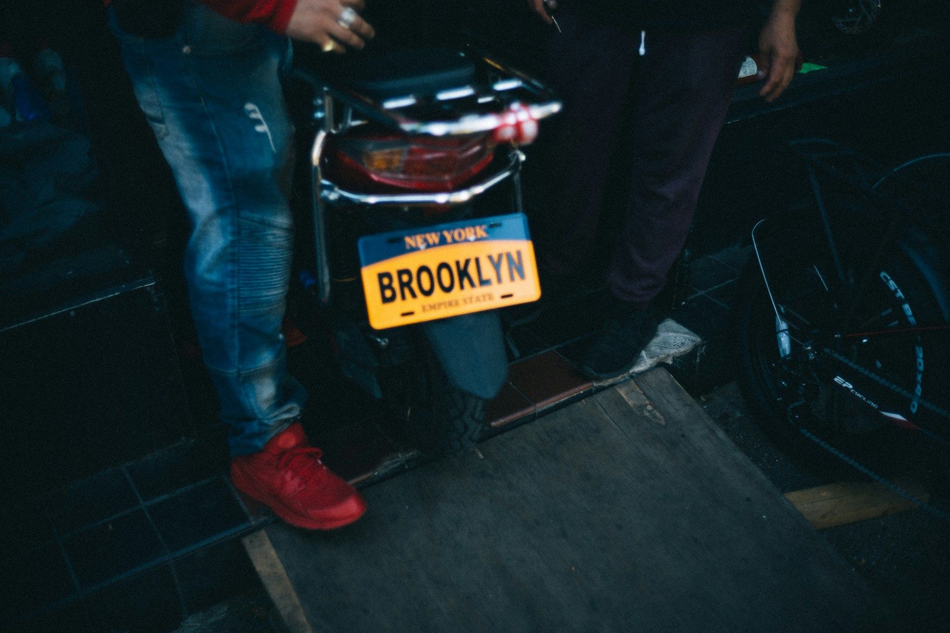 Motorcycle with a New York license plate reading Brooklyn parked indoors with two people standing beside it.