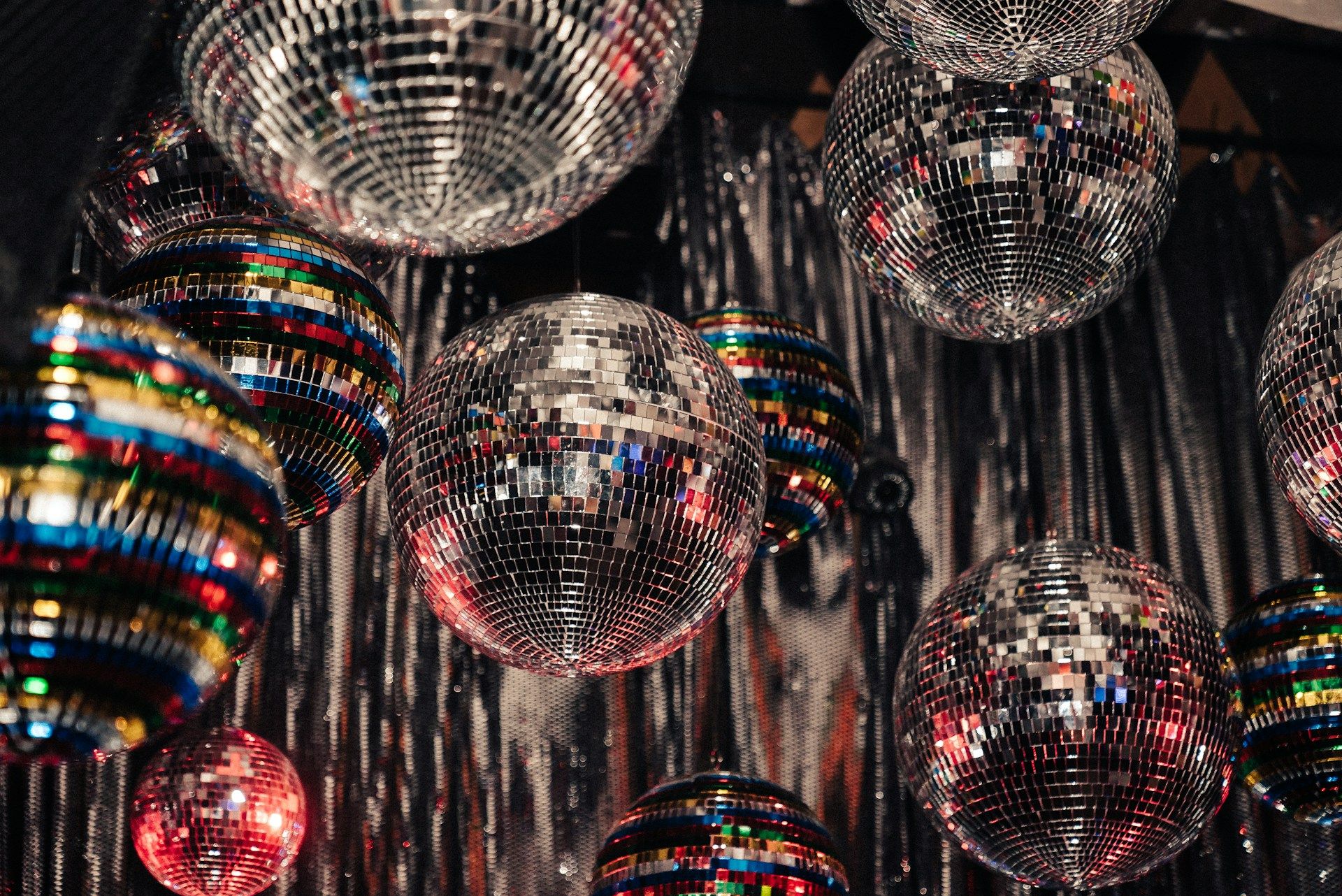 Collection of colorful mirrored disco balls hanging from a ceiling against a shimmering backdrop.