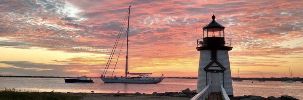 Lighthouse in Nantucket, Massachusetts overlooking sailboats on calm water at sunset with colorful clouds in the sky.