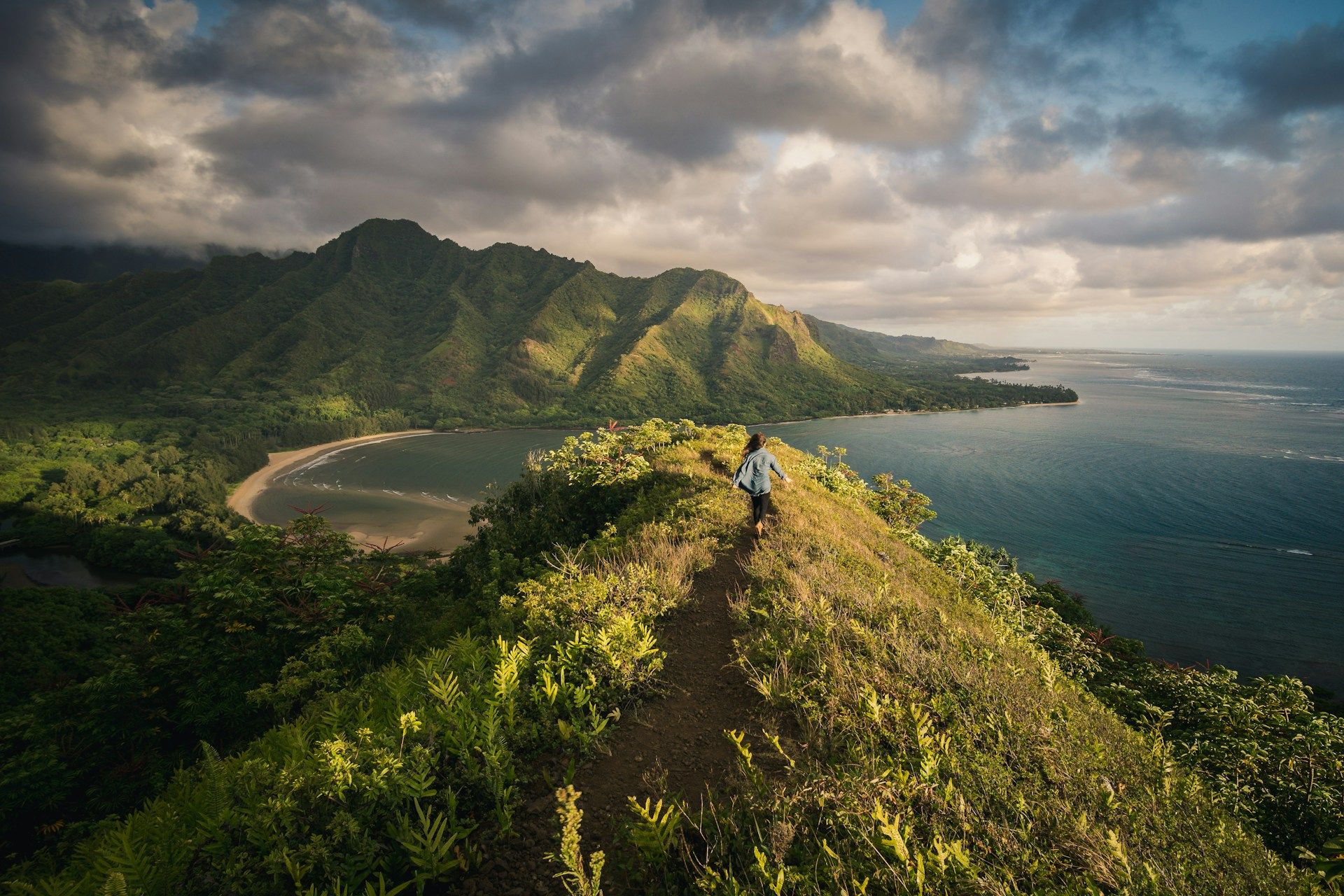 Person hiking along a scenic coastal ridge trail overlooking lush green mountains and the ocean.