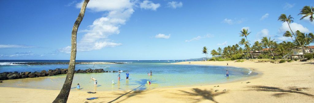 Sunny day at Poipu Beach, Hawaii, with palm trees, golden sand, and people swimming in clear blue water.