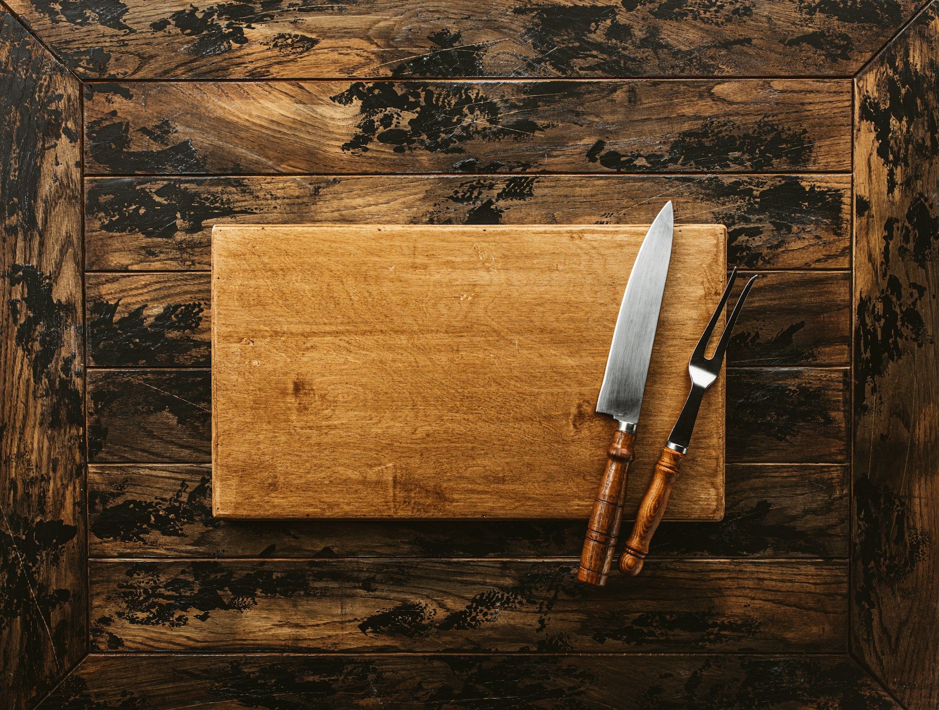 Wood-handled chef's knife and carving fork resting on a wooden cutting board over a rustic wood table.