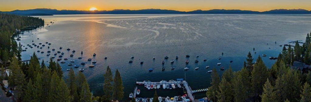 Aerial view of Lake Tahoe at sunset with boats anchored near the shore and a small marina surrounded by trees.