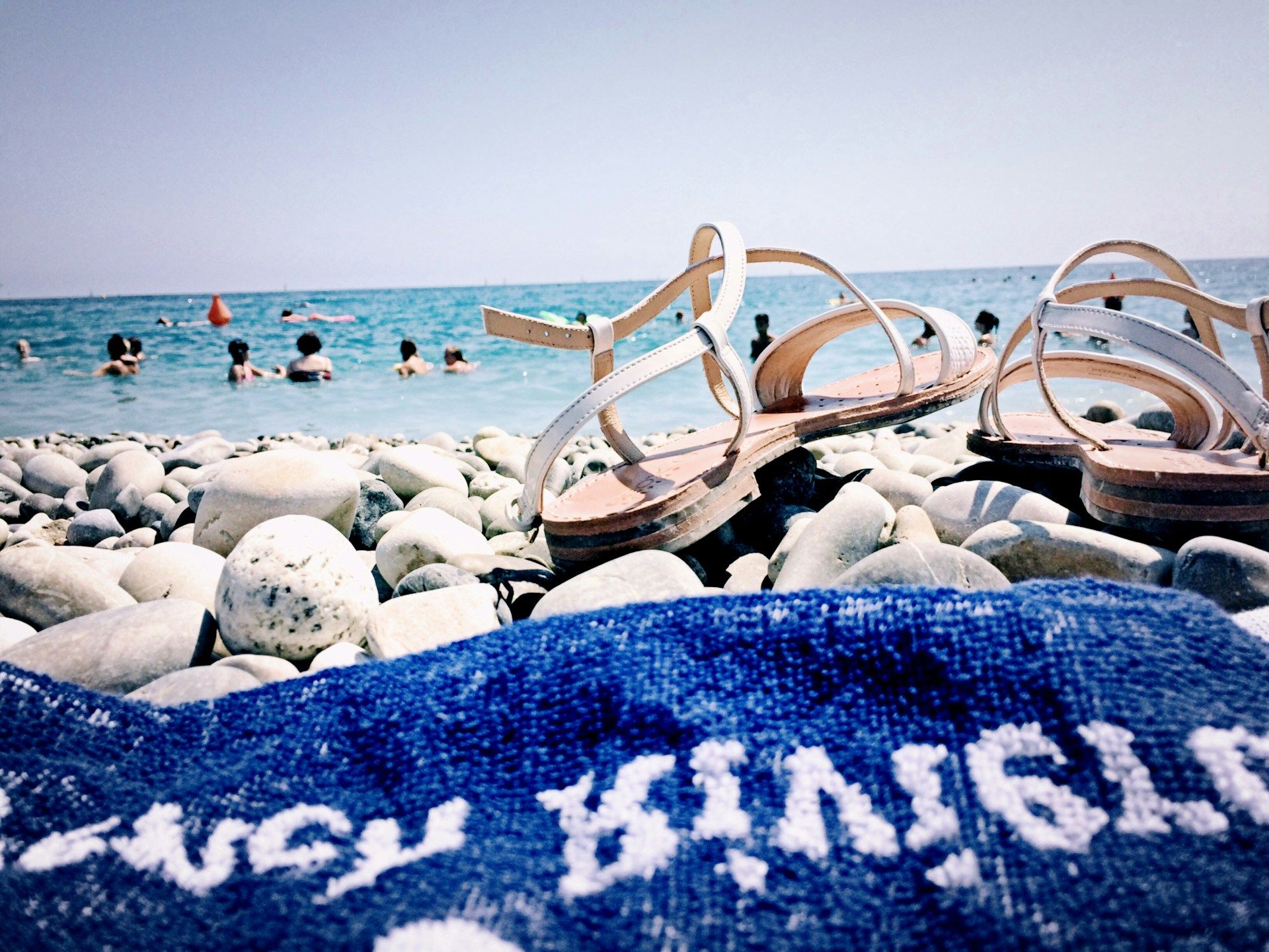 Sandals resting on a monogrammed beach towel on a pebble beach by the sea.